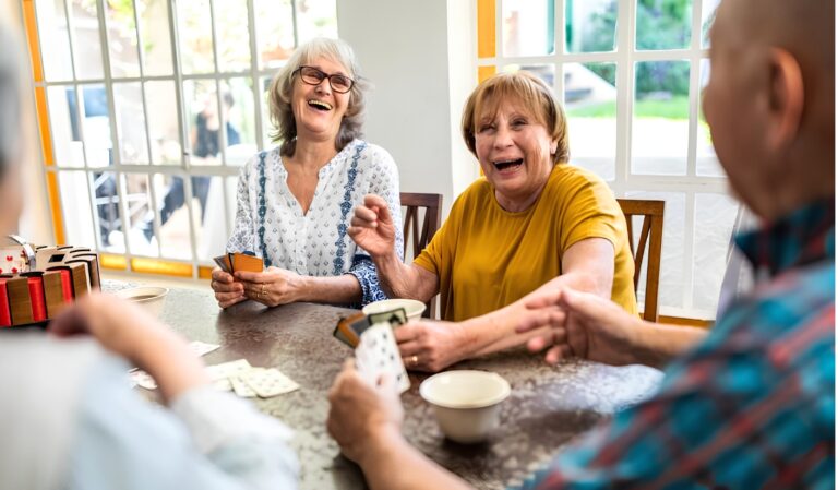 Group of smiling seniors playing cards at a table, enjoying a social moment indoors.