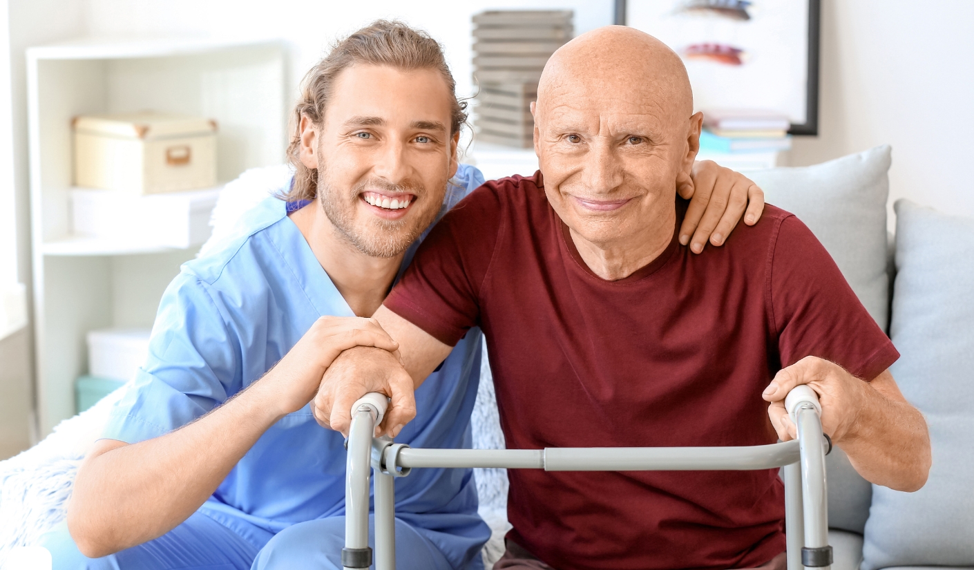 Caregiver in blue scrubs helping an elderly man with a walker, both smiling in a living room.