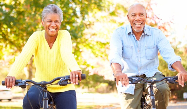 Two smiling adults ride bikes together on a sunny park path.