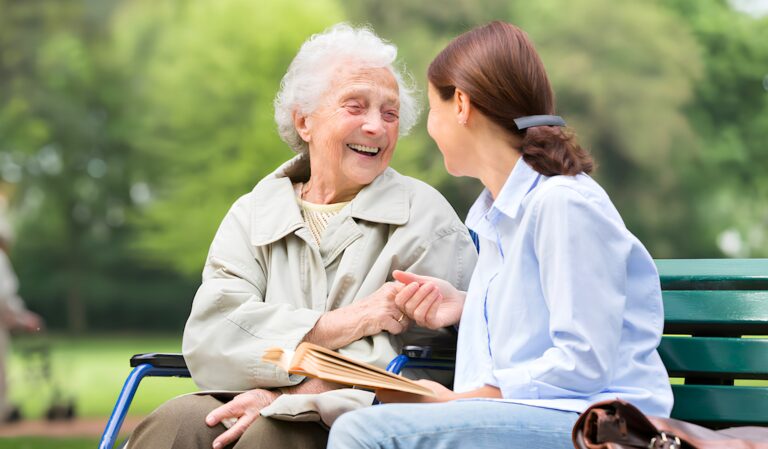 Elderly woman in a wheelchair on a park bench, smiling while holding hands with a caregiver outdoors.